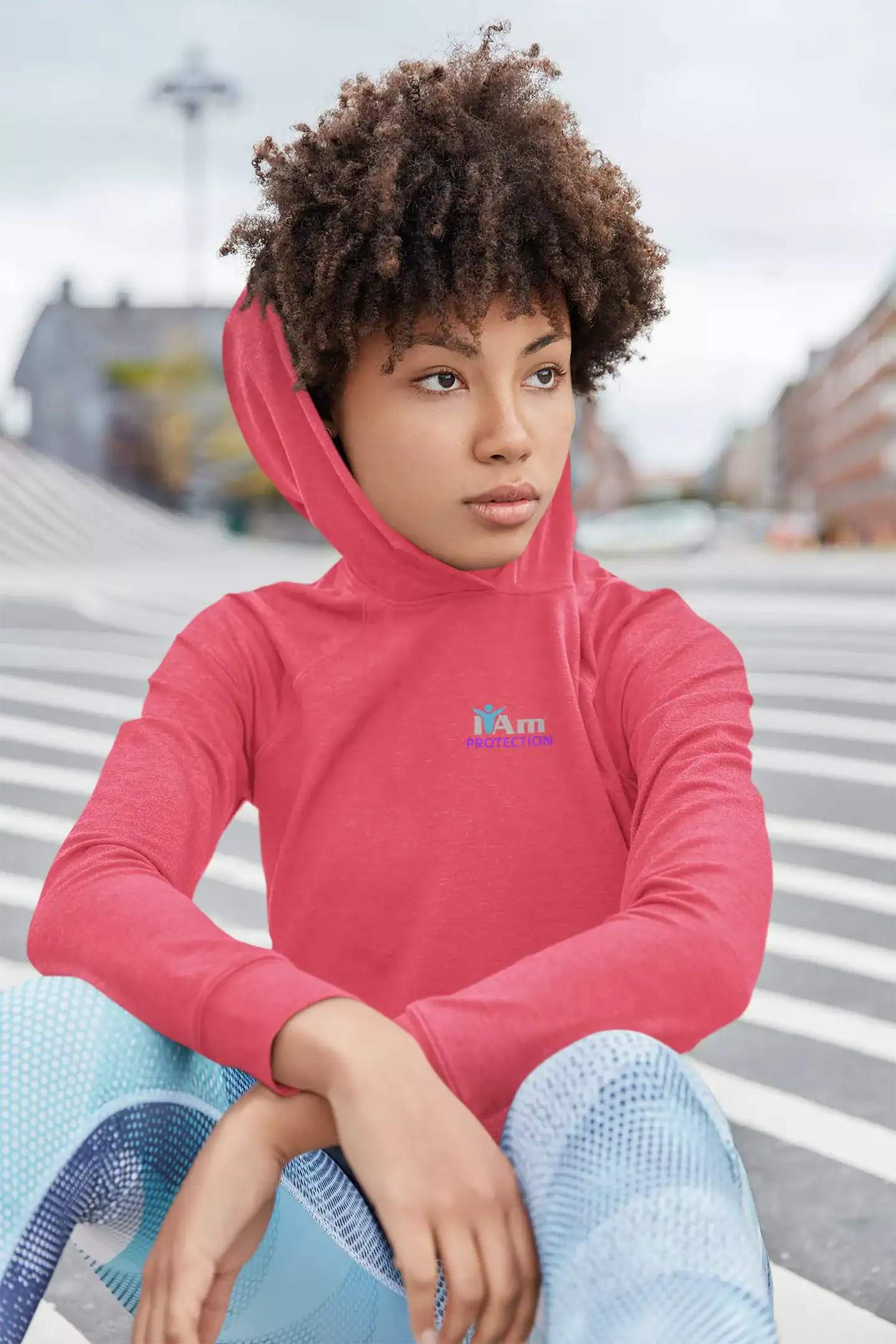 Young woman in pink hoodie sitting outdoors on city street.