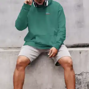 Young man in green hoodie, black cap, headphones, sitting against concrete wall.