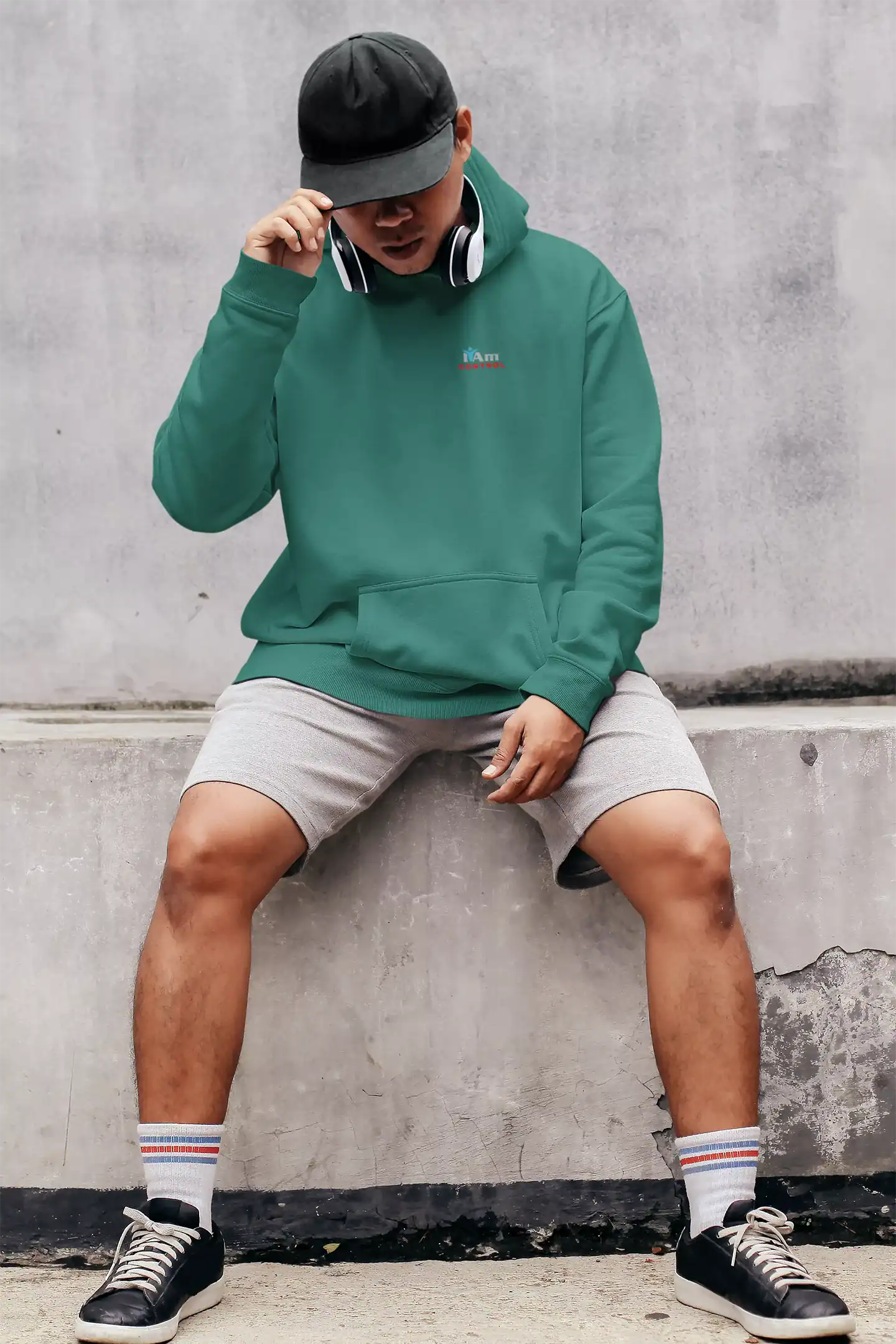Young man in green hoodie, black cap, headphones, sitting against concrete wall.