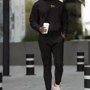 Woman in black hoodie and joggers walking outdoors with coffee.