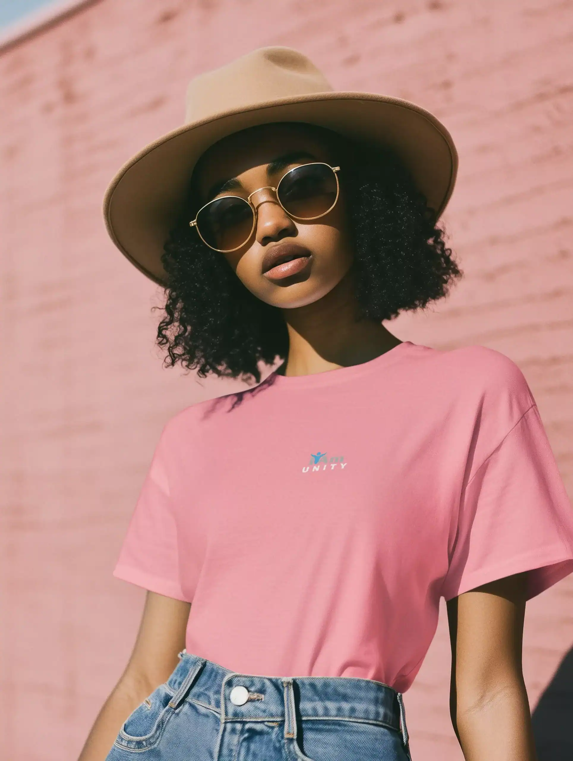 Stylish woman wearing pink 'I Am Unity' graphic tee, sunglasses, and hat outdoors.